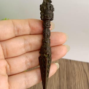 May include: A detailed bronze-colored ritual dagger, featuring intricate carvings and a pointed blade. The handle has a detailed figure at the top and a series of decorative bands. The dagger is held in a hand.