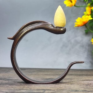 May include: A bronze incense holder with a curved, leaf-like design. A yellow incense cone is placed on top. The holder is on a wooden surface, with yellow flowers in the background.
