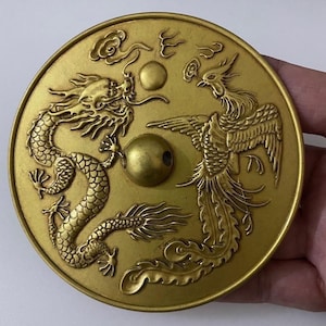Puede incluir: Disco redondo de metal dorado antiguo con detallados grabados en relieve de un dragón y un fénix enfrentados. El disco tiene una esfera central y es probablemente un objeto decorativo o una obra de arte.