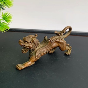 May include: A bronze-colored metal figurine of a mythical creature resembling a winged lion or dragon. The creature is depicted in a pouncing stance with its mouth open, showing teeth, and features detailed scales, wings, and a curved tail.