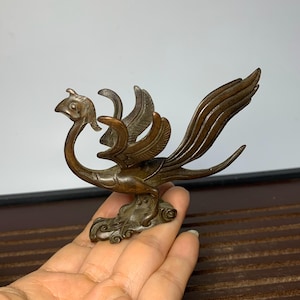 Op de afbeelding: Een bronzen phoenix sculptuur met uitgestrekte vleugels en een lange, vloeiende staart. De vogel zit op een wolkenachtige basis met wervelende details. De sculptuur heeft ingewikkelde veerdetaillering.