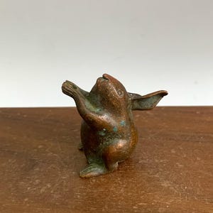 May include: A small bronze rabbit figurine with outstretched arms. The rabbit has long ears and a weathered appearance, with green patina. The figurine is set on a wooden surface. The rabbit is approximately 7 cm tall.