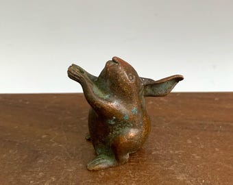 May include: A small, bronze figurine of a rabbit with its arms raised. The rabbit has long ears and a slightly weathered appearance, with hints of green patina. The figurine is set against a wooden surface.