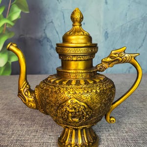 May include: A gold-coloured, ornate teapot with a dragon-shaped handle and spout. The teapot features intricate embossed patterns and a decorative lid. The base is a pedestal design. The teapot is made of metal.