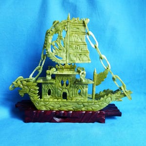 Puede incluir: Una detallada escultura de barco de jade verde con intrincadas tallas, incluyendo una cabeza de dragón en la proa y una vela decorativa. El barco está montado sobre una base de madera roja oscura, sobre un fondo azul.