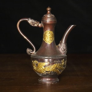 May include: A bronze-colored decorative pitcher with gold accents. The pitcher features a curved handle with a dragon head, a spout, and ornate gold phoenix and dragon designs. The lid is topped with a small finial.