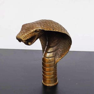 Könnte beinhalten: Eine detaillierte Bronzeskulptur einer Kobra mit erhobener Haube, der Kopf ist nach links gerichtet. Die Skulptur ist auf einem runden Sockel montiert.