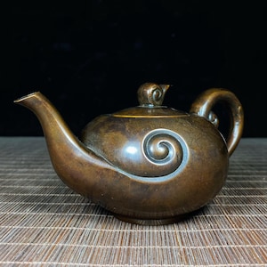 May include: A bronze-colored teapot with a curved spout and handle. The teapot features a spiral design on its side and a small, decorative lid. The teapot is displayed on a woven mat, with a dark background.