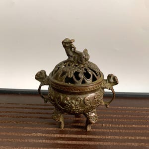 Op de afbeelding: Een kleine, bronzen wierookbrander met ingewikkelde bloemmotieven en een deksel in de vorm van een draak. De brander heeft drie poten en twee handvatten.
