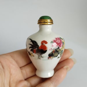 May include: A small, white ceramic bottle with a green and gold lid. The bottle is decorated with a rooster and floral design in red, black, and yellow. The bottle is held in a hand.