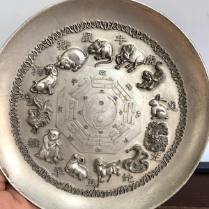 May include: A silver-coloured plate with the Chinese zodiac animals encircling a Yin and Yang symbol. The plate has a decorative border and detailed relief carvings of each animal, including a rat, ox, tiger, rabbit, dragon, snake, horse, sheep, monkey, rooster, dog, and pig.