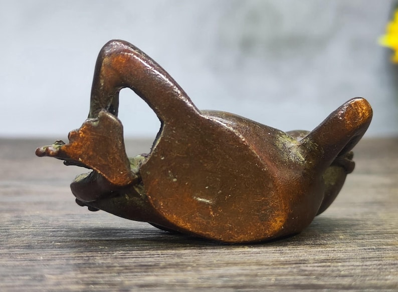 May include: A bronze frog figurine in a relaxed pose, with its legs extended. The frog is lying on its back, with its arms and legs in the air. The figurine has a smooth, polished surface and is a warm brown color.