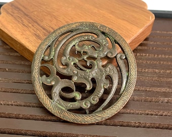 Puede incluir: Un colgante circular de color bronce con un diseño intrincado de un dragón en un patrón de espiral. El colgante tiene un borde texturizado y está ligeramente desgastado.