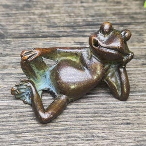 May include: A bronze frog figurine in a relaxed pose, resting its chin on its hand. The frog's body has a smooth, polished finish with hints of blue and green. The figurine is on a wooden surface.