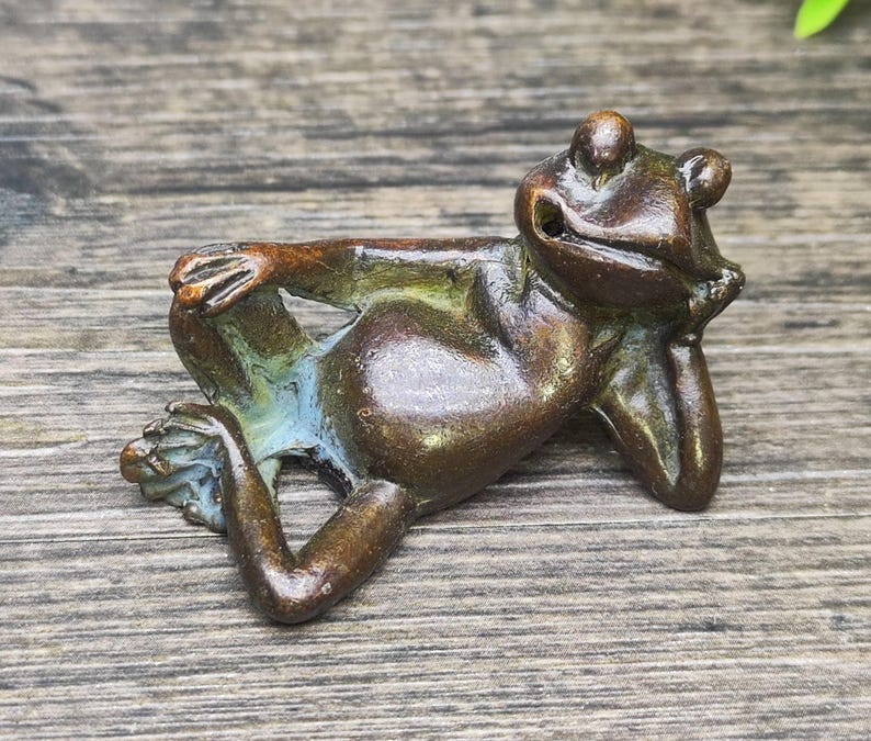 May include: A bronze frog figurine in a relaxed pose, resting its chin on its hand. The frog's body has a smooth, polished finish with hints of blue and green. The figurine is on a wooden surface.