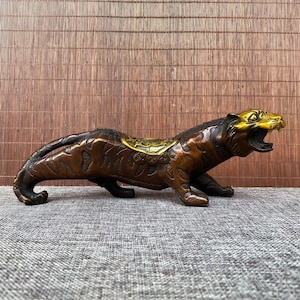 May include: Bronze tiger sculpture with a gold-colored head and saddle. The tiger is in a crouching position with its mouth open, revealing its teeth. The sculpture has intricate details and a textured surface, showcasing the craftsmanship.