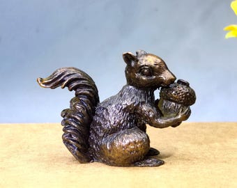 May include: A bronze squirrel figurine holding an acorn. The squirrel has detailed fur texture and a bushy tail. The acorn is held in its paws and mouth. The figurine is a decorative item, likely for a home or office.