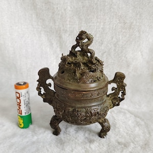 May include: Bronze incense burner with intricate dragon and floral detailing. The burner has three legs and two handles. A standard AA battery is next to the burner for size comparison.