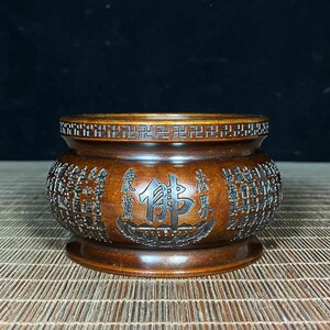 May include: A brown, round incense burner with intricate carvings and Chinese characters. The burner features a decorative rim and a base. The word "Buddha" is prominently displayed in the center. The overall design is detailed and ornate.