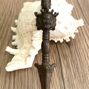 May include: A bronze dagger with detailed carvings, including a face at the hilt and a pointed blade. The dagger is set against a seashell and a wooden surface. The dagger's length is approximately 15 cm.