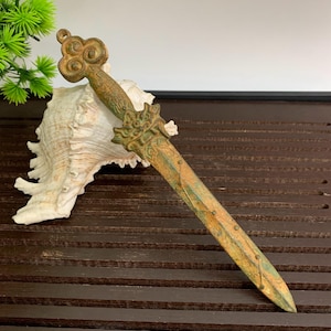 Könnte beinhalten: Ein antiker Bronzedolch mit aufwendigen floralen und geometrischen Mustern. Die Klinge hat ein patiniertes, gealtertes Aussehen, und der Griff weist eine dekorative Schlaufe auf. Der Dolch liegt auf einer Holzoberfläche.