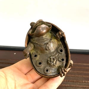 Puede incluir: Una escultura de bronce de una rana sentada dentro de un caparazón de tortuga. La rana lleva un collar con un colgante. El caparazón de la tortuga tiene un diseño floral y varios agujeros.