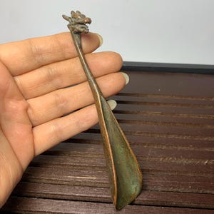 Könnte beinhalten: Ein bronzefarbener Schuhanzieher mit einem Drachenkopf-Design. Der Schuhanzieher hat einen langen, schlanken Griff und eine breite, gebogene Klinge. Der Drachenkopf ist mit Schuppen und einem grimmigen Ausdruck versehen. Das Metall hat eine Patina.