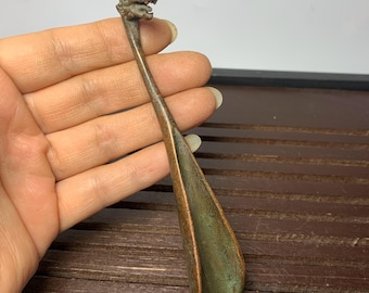 May include: A bronze-colored shoehorn with a dragon head design. The shoehorn has a long, slender handle and a wide, curved blade. The dragon head is detailed with scales and a fierce expression. The metal has a patina, giving it an antique appearance.