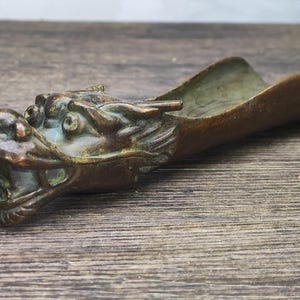May include: A bronze tea scoop featuring a dragon head. The scoop has a curved, trough-like form and a detailed dragon head at the handle end. The dragon's mouth is open, and the scoop has a patina finish, giving it an aged appearance.