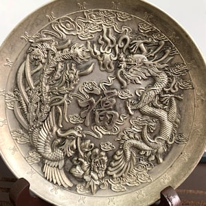 Könnte beinhalten: Ein silberfarbener Zierteller mit aufwendigen Reliefschnitzereien. Der Teller zeigt ein zentrales chinesisches Schriftzeichen, umgeben von Drachen, Phönixen und Wolkenmotiven. Der Teller wird auf einem Holzständer präsentiert.