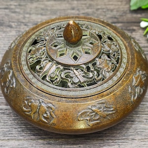 May include: A bronze-colored incense burner with intricate relief carvings. The round burner features a detailed lid with geometric and floral cutouts, and a small, cone-shaped handle. The body of the burner is adorned with relief patterns.
