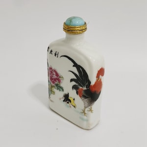 May include: A white ceramic bottle with a rooster and floral design. The bottle has a turquoise stopper with a gold rim. The rooster is red, black, and yellow. The bottle has Chinese characters on the side.