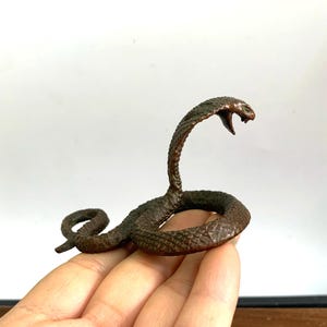 Op de afbeelding: Een klein bronzen beeldje van een cobra met zijn kap op, de slang is opgerold in een verdedigende houding.