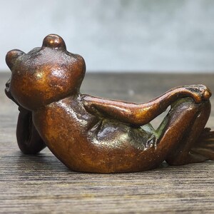 May include: A bronze frog figurine in a relaxed pose, resting on its back with arms crossed. The frog has a smooth, polished surface and is detailed with visible toes and facial features. The figurine is set against a blurred background.