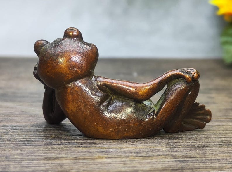 May include: A bronze frog figurine in a relaxed pose, resting on its back with arms crossed. The frog has a smooth, polished surface and is detailed with visible toes and facial features. The figurine is set against a blurred background.