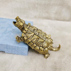 May include: A detailed, antique-style brass figurine of an Ankylosaurus dinosaur. The sculpture features textured scales, a spiked back, and an open mouth, showcasing intricate craftsmanship. The dinosaur is positioned on a light blue surface.