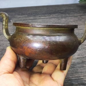 May include: A small, antique bronze incense burner with three legs and two curved handles. The vessel has a rounded body and a dark, aged patina, suggesting its use over time. The design is simple yet elegant.