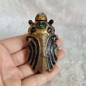 May include: A detailed, antique-style decorative bottle. The bottle is shaped like an insect, with a gold-colored body and black and blue accents. The top of the bottle has a small, clear stopper. The bottle is held in a hand.