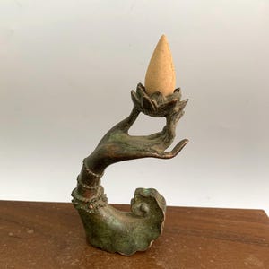 Può includere: Una scultura di una mano in bronzo che tiene un bruciatore di incenso a forma di fiore di loto. Il cono di incenso è beige e si trova al centro del fiore di loto.