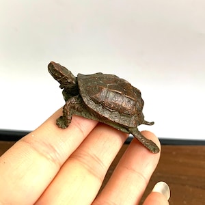 Pode incluir: Uma pequena figura de tartaruga de bronze com uma casca e pernas detalhadas. A tartaruga tem aproximadamente 5 cm de comprimento e está em posição de caminhar.