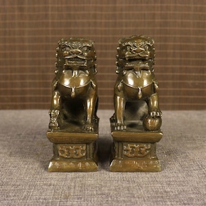 May include: Two bronze-colored Foo Dog statues. Each statue is in a seated position on a decorative base. The Foo Dogs have detailed features, including a mane, a stern expression, and a ball under one paw. The statues are displayed against a neutral background.