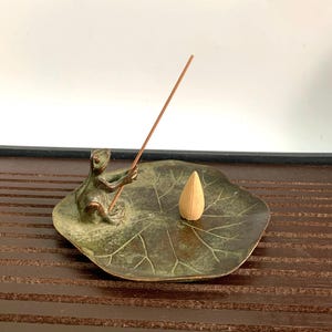 Puede incluir: Un quemador de incienso de bronce con forma de rana sentado en una hoja de loto. El quemador de incienso tiene un pequeño agujero en el centro para un cono de incienso. La rana sostiene un palo de incienso.