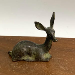 Può includere: Una piccola scultura in bronzo di un cervo sdraiato. Il cervo è di colore verde scuro con una finitura liscia e lucida.