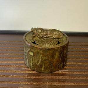 Puede incluir: Un quemador de incienso de bronce con un diseño de cigarra en la tapa. El quemador es cilíndrico y tiene una superficie texturizada que se asemeja a la corteza de un árbol.