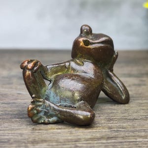 May include: A bronze frog figurine in a relaxed pose, resting its head on its hand. The frog has a weathered appearance with hints of green and blue patina. The sculpture is detailed and appears to be made of metal.