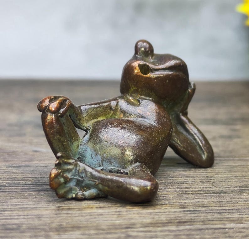 May include: A bronze frog figurine in a relaxed pose, resting its head on its hand. The frog has a weathered appearance with hints of green and blue patina. The sculpture is detailed and appears to be made of metal.