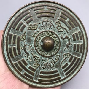 Op de afbeelding: Een ronde, antieke bronzen spiegel met ingewikkelde reliëfsnijwerk. De spiegel heeft een centrale knop en is versierd met dierenfiguren en geometrische patronen. Het brons heeft een verweerde uitstraling, met groene patina.