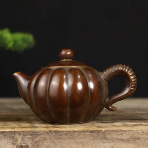 Puede incluir: Una tetera marrón con forma de calabaza, con un asa curva y una tapa pequeña. La tetera está hecha de madera oscura y se encuentra sobre una superficie de madera. El asa tiene un diseño texturizado.