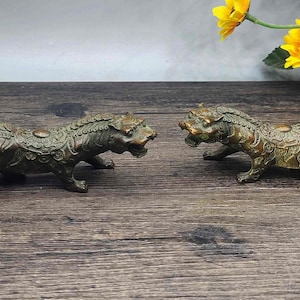 Può includere: Due statuette di tigre in bronzo, una di fronte all'altra, con incisioni dettagliate. Le tigri sono in posizione accovacciata, con la bocca aperta. Le statuette sono esposte su una superficie di legno scuro.