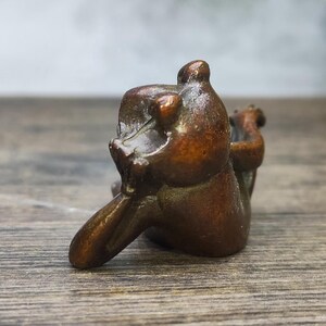 May include: A small, bronze-colored sculpture of a bear in a seated position. The bear is depicted with its arms raised and its head tilted back. The sculpture has a smooth, polished finish and appears to be made of metal.